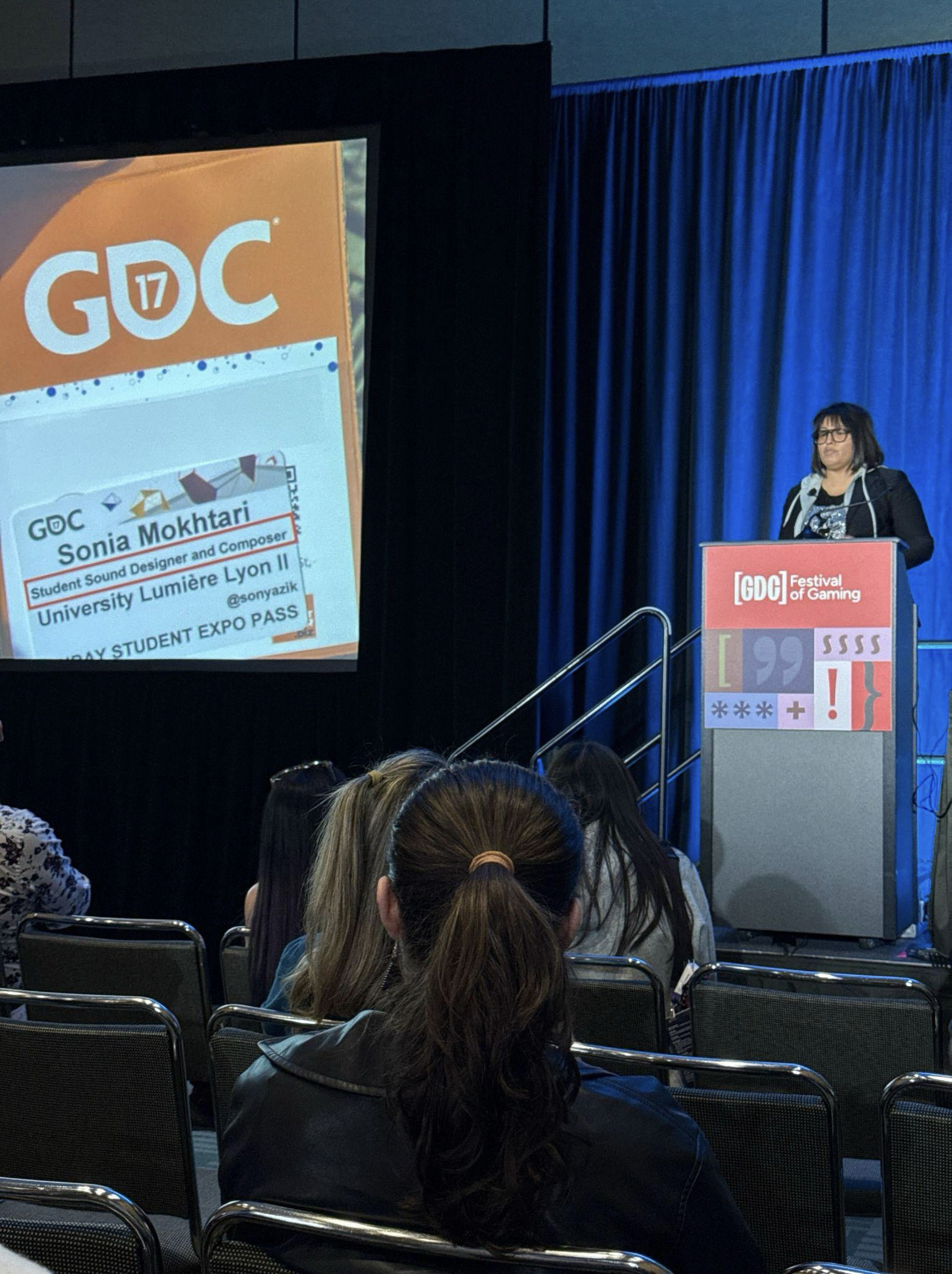 A presentation at the GDC Festival of Gaming featuring a slide of a student attendee badge on a large screen while Sonia stands at the podium.